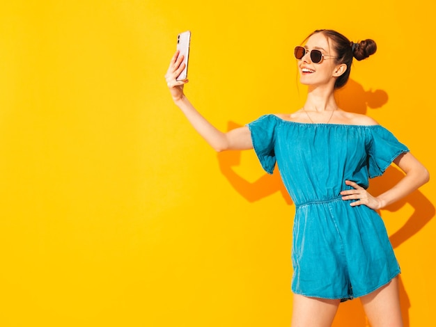 Young beautiful smiling female in trendy summer jeans overalls Sexy carefree woman with two horns hairstyle posing near yellow wall in studio Positive model having fun Isolated Taking selfie