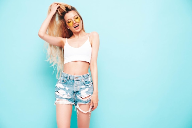 Young beautiful smiling female in trendy summer jeans shorts Sexy carefree blond woman posing near blue wall in studio Positive model having fun Cheerful and happy in sunglasses Isolated