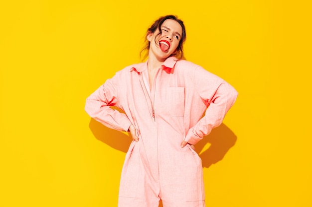 Young beautiful smiling female in trendy summer pink overalls Sexy carefree woman posing near yellow wall in studio Positive model having fun and going crazy Cheerful and happy Shows tongue