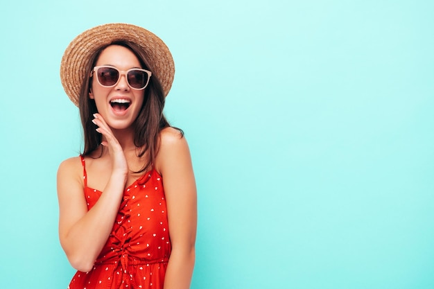 Young beautiful smiling female in trendy summer red dress Sexy carefree woman posing near blue wall in studio Positive brunette model having fun in hat and sunglasses Screams and shouts