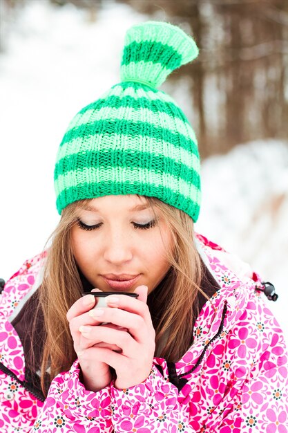 Young beautiful smiling girl in a crimson jacket and green hat drinking hot tea from a thermos in snowy mountains