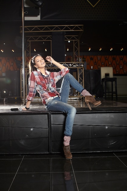 Free photo young beautiful smiling pop star singer with microphone sitting on the scene in the club