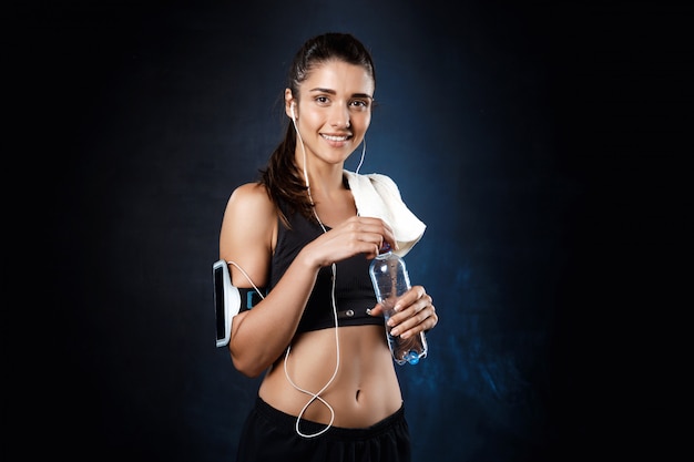 Young beautiful sportive girl holding water over dark wall.