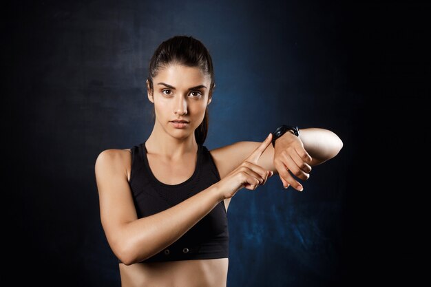 Young beautiful sportive girl pointing at watch over dark wall.