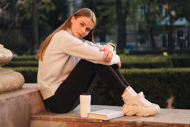 Young beautiful stylish student girl in cozy sweater thoughtfully looking in camera while studying outdoor