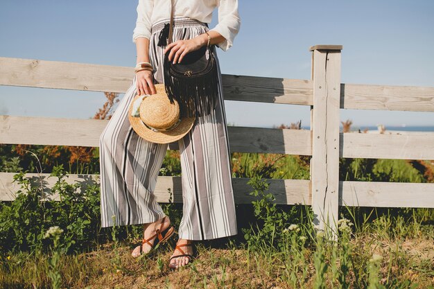 Young beautiful stylish woman, spring summer fashion trend, boho style, straw hat, countryside weekend, sunny, smiling, fun, sunglasses, black purse, striped trousers, details, accessories