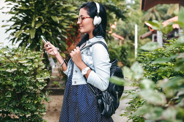 Free photo young beautiful stylish woman using smartphone, headphones, eye glasses, summer, vintage denim outfit, smiling, happy, positive