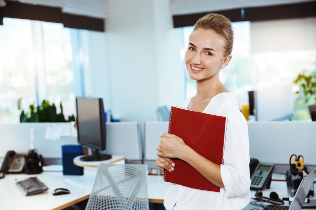 Young beautiful successful businesswoman smiling, posing, holding folder, over office