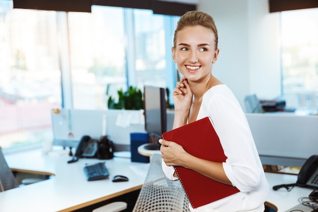 Free photo young beautiful successful businesswoman smiling, posing, holding folder, over office