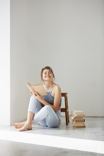 Young beautiful tender woman smiling holding book sitting on floor over white wall early in morning.