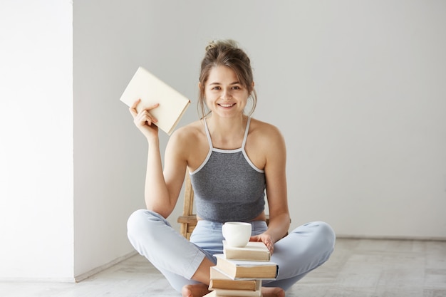 Free photo young beautiful tender woman smiling holding book sitting on floor over white wall early in morning.