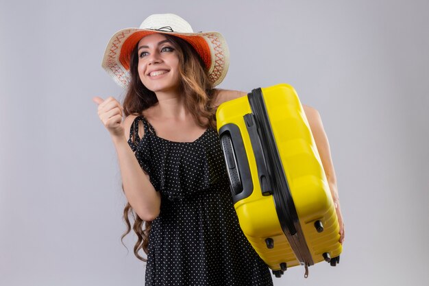 Young beautiful traveler girl in dress in polka dot in summer hat holding suitcase looking exited and happy showing thumbs up rejoicing her success and victory standing over white background