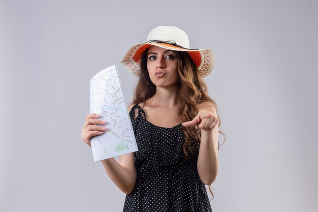 Young beautiful traveler girl in dress in polka dot in summer hat pointing with finger to camera with skeptic expression on face standing over white background