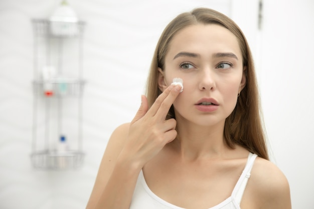 Young beautiful woman applying cream to face in the bathroom