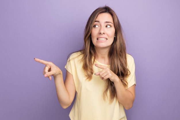 Young beautiful woman in beige t-shirt looking aside confused pointing with index figners to the side standing over purple