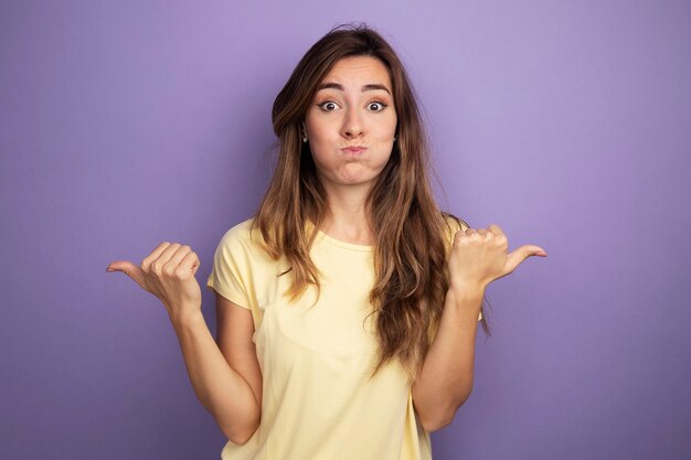 Young beautiful woman in beige t-shirt looking at camera confused blowing cheeks pointing with thumbs to the different sides 