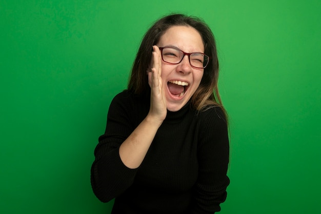 Free Photo young beautiful woman in a black turtleneck and glasses happy and excited looking at front shouting with hand near mouth standing over green wall