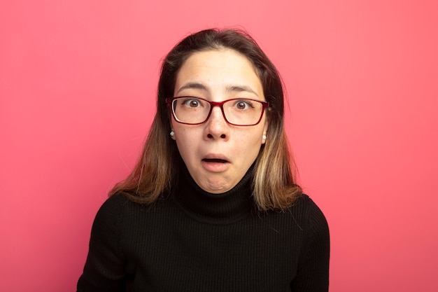 Free Photo young beautiful woman in a black turtleneck and glasses looking at front being confused standing over pink wall