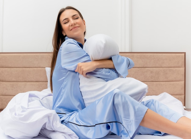 Young beautiful woman in blue pajamas sitting on bed with pillow happy and positive looking joyful smiling in bedroom interior on light background