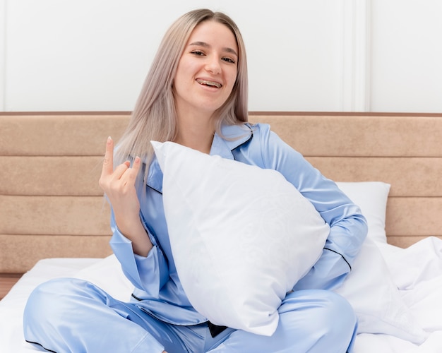 Free photo young beautiful woman in blue pajamas sitting on bed with pillow looking at camera smiling with happy face resting showing rock symbol in bedroom interior on light background