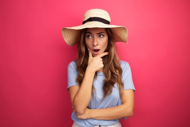 Young beautiful woman in blue t-shirt and summer hat looking aside surprised standing over pink background