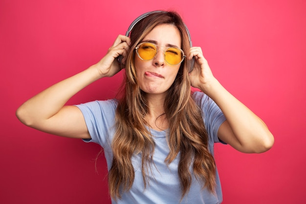 Young beautiful woman in blue t-shirt wearing yellow glasses with headphones looking aside happy and joyful sticking out tongue standing over pink background