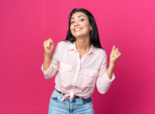 Young beautiful woman in casual clothes happy and excited clenching fists standing on pink