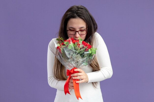 young beautiful woman in casual clothes holding bouquet of red roses happy and cheerful smiling valentines day concept standing over purple wall