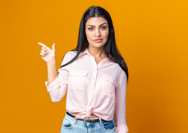 Young beautiful woman in casual clothes looking at front with confident expression pointing with index finger to the side standing over orange wall