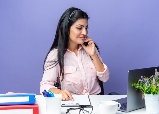 Young beautiful woman in casual clothes wearing headset with microphone looking confident smiling sitting at the table with laptop over blue background working in office