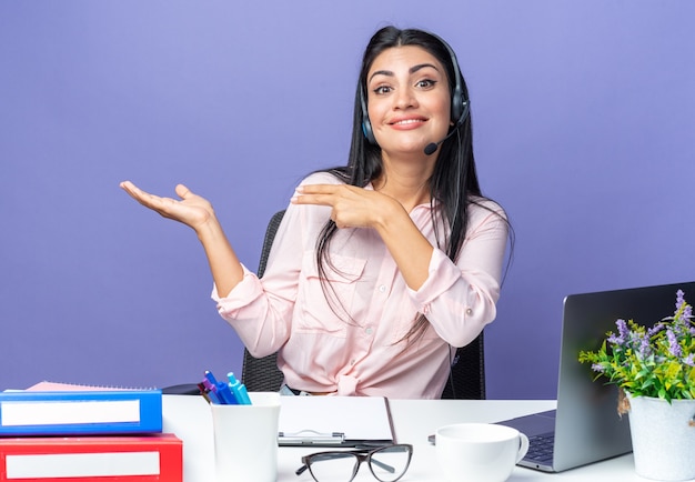 Free photo young beautiful woman in casual clothes wearing headset with microphone smiling presenting with arm of hand sitting at the table with laptop over blue wall working in office