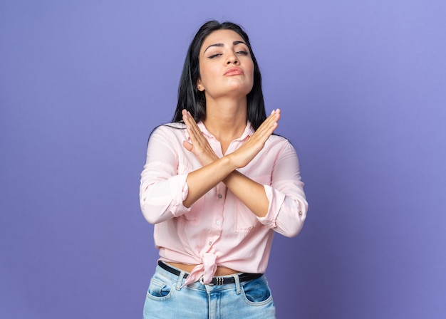 Young beautiful woman in casual clothes with serious face making stop gesture crossing hands standing on blue