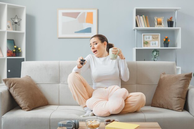 Young beautiful woman in casual clothing sitting on a couch at home interior holding remote and a cup of tea looking scared and worried spending weekend at home