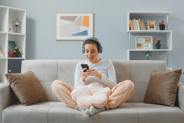 Young beautiful woman in casual clothing with headphones sitting on a couch at home interior holding smartphone happy and surprised looking at the screen of her mobile smiling cheerfully relaxing