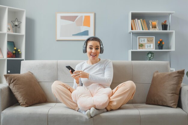 Young beautiful woman in casual clothing with headphones sitting on a couch at home interior holding smartphone looking at camera happy and positive smiling broadly relaxing at home