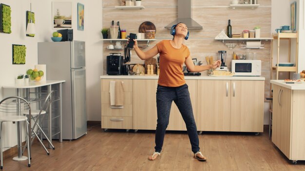 Free photo young beautiful woman dancing while listening music in blue wirelless headphones in the kitchen. energetic, positive, happy, funny and cute housewife dancing alone in the house. entertainment and leiu