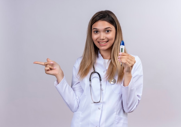 Free photo young beautiful woman doctor wearing white coat with stethoscope holding digital thermometer smiling cheerfully pointing with index finger to the side