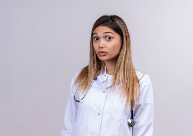 Young beautiful woman doctor wearing white coat with stethoscope looking amazed and surprised