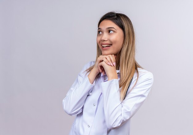 Young beautiful woman doctor wearing white coat with stethoscope looking aside holding hands together exited waiting for surprise