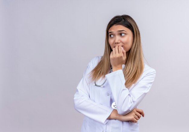 Young beautiful woman doctor wearing white coat with stethoscope looking aside stressed and nervous biting nails