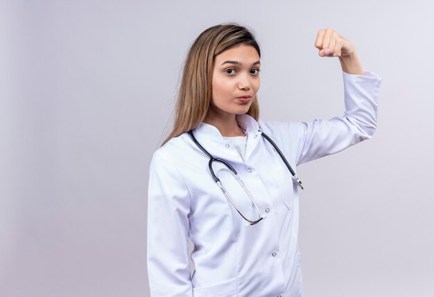 Young beautiful woman doctor wearing white coat with stethoscope looking confident raising fist posing like a winner