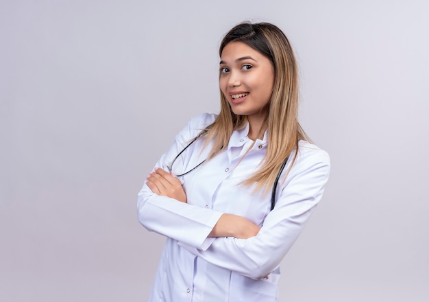 Young beautiful woman doctor wearing white coat with stethoscope looking confident standing with arms crossed