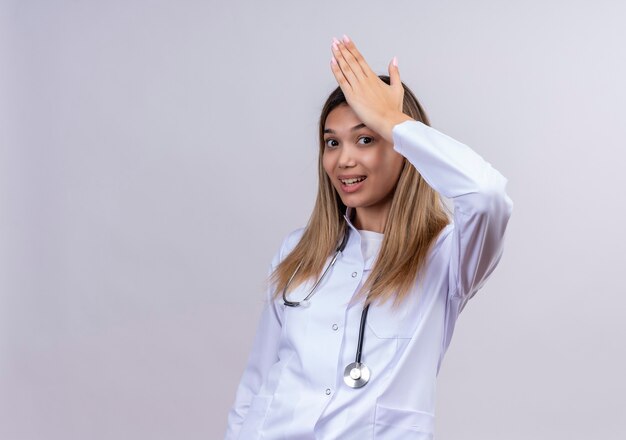 Young beautiful woman doctor wearing white coat with stethoscope looking confused and uncertain with hand on head for mistake