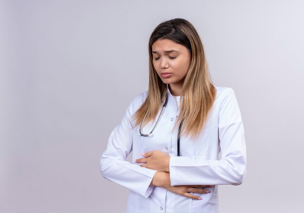 Young beautiful woman doctor wearing white coat with stethoscope looking unwell touching her belly suffering from pain