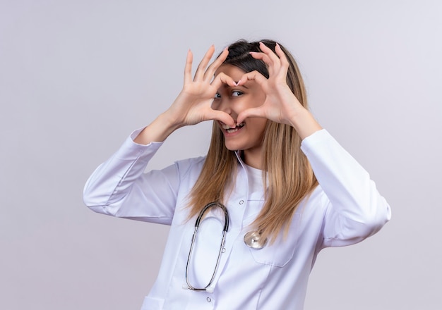 Free Photo young beautiful woman doctor wearing white coat with stethoscope making romantic heart gesture with fingers looking through this sign being lovely