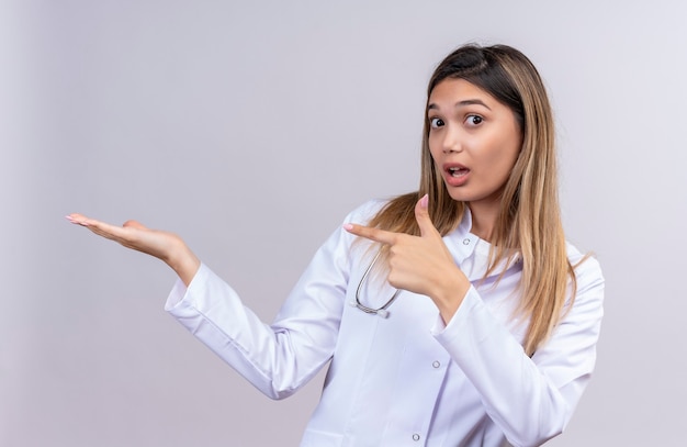 Young beautiful woman doctor wearing white coat with stethoscope presenting with her arm of her hand and pointing with finger to the side looking surprised