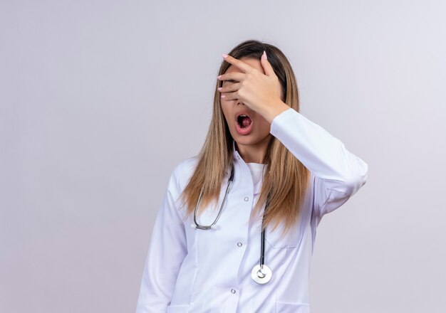 Young beautiful woman doctor wearing white coat with stethoscope shocked covering eyes with hand