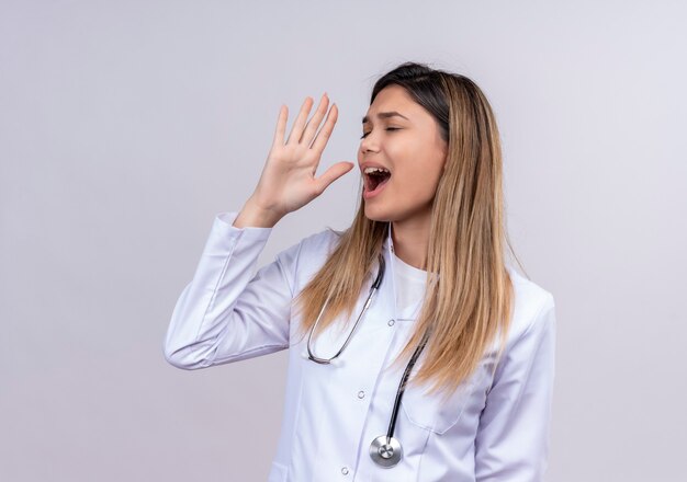 Young beautiful woman doctor wearing white coat with stethoscope shouting with hand near mouth