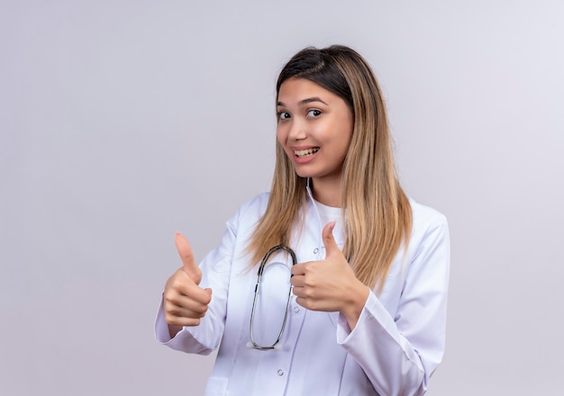 Young beautiful woman doctor wearing white coat with stethoscope smiling cheerfully showing thumbs up