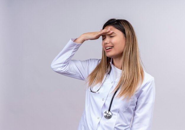 Young beautiful woman doctor wearing white coat with stethoscope standing with hand on head for mistake looking confused